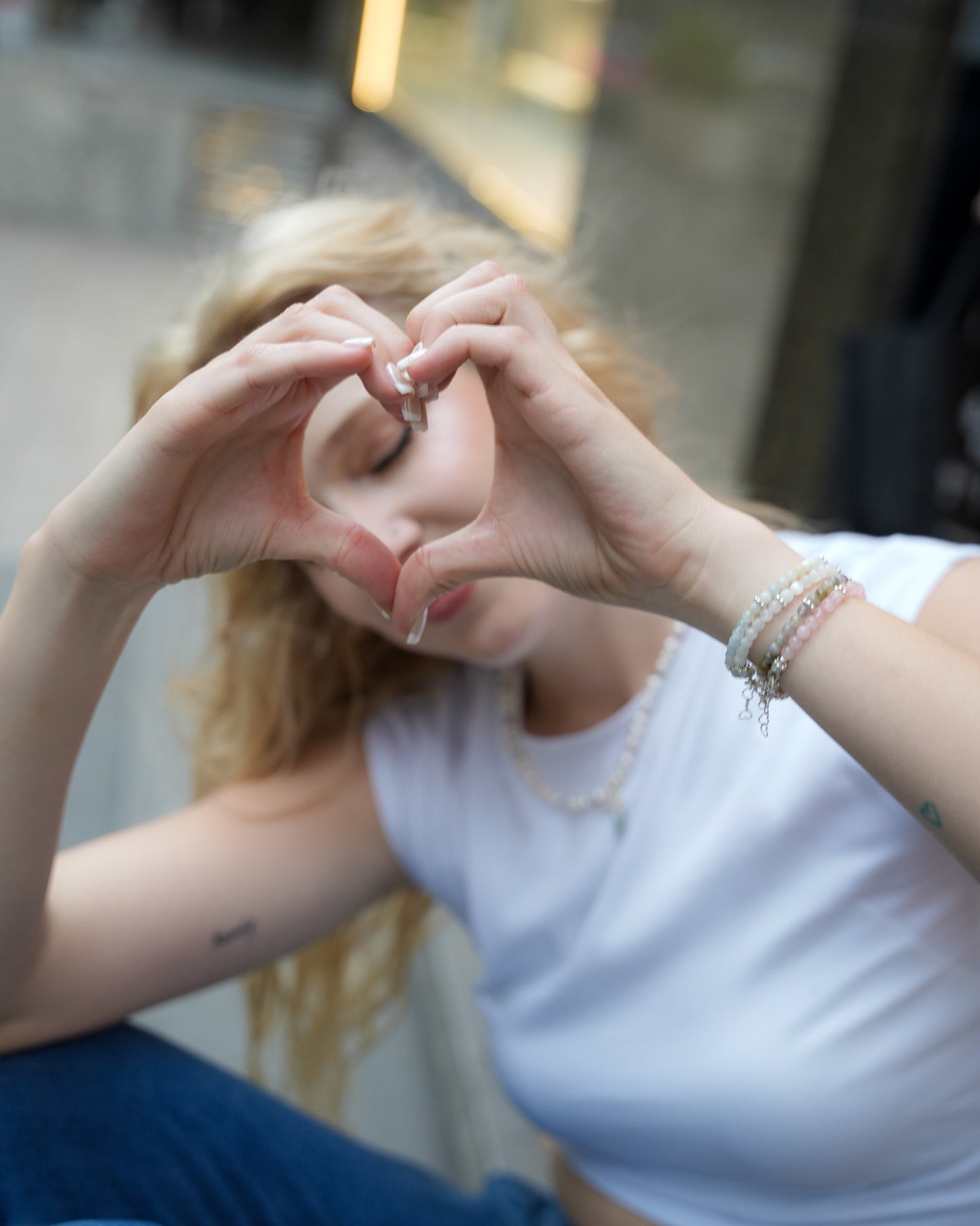 Woman forming a heart with her hands while wearing layered gemstone and silver bracelets and a pearl necklace – casual, playful jewelry moment by elswre