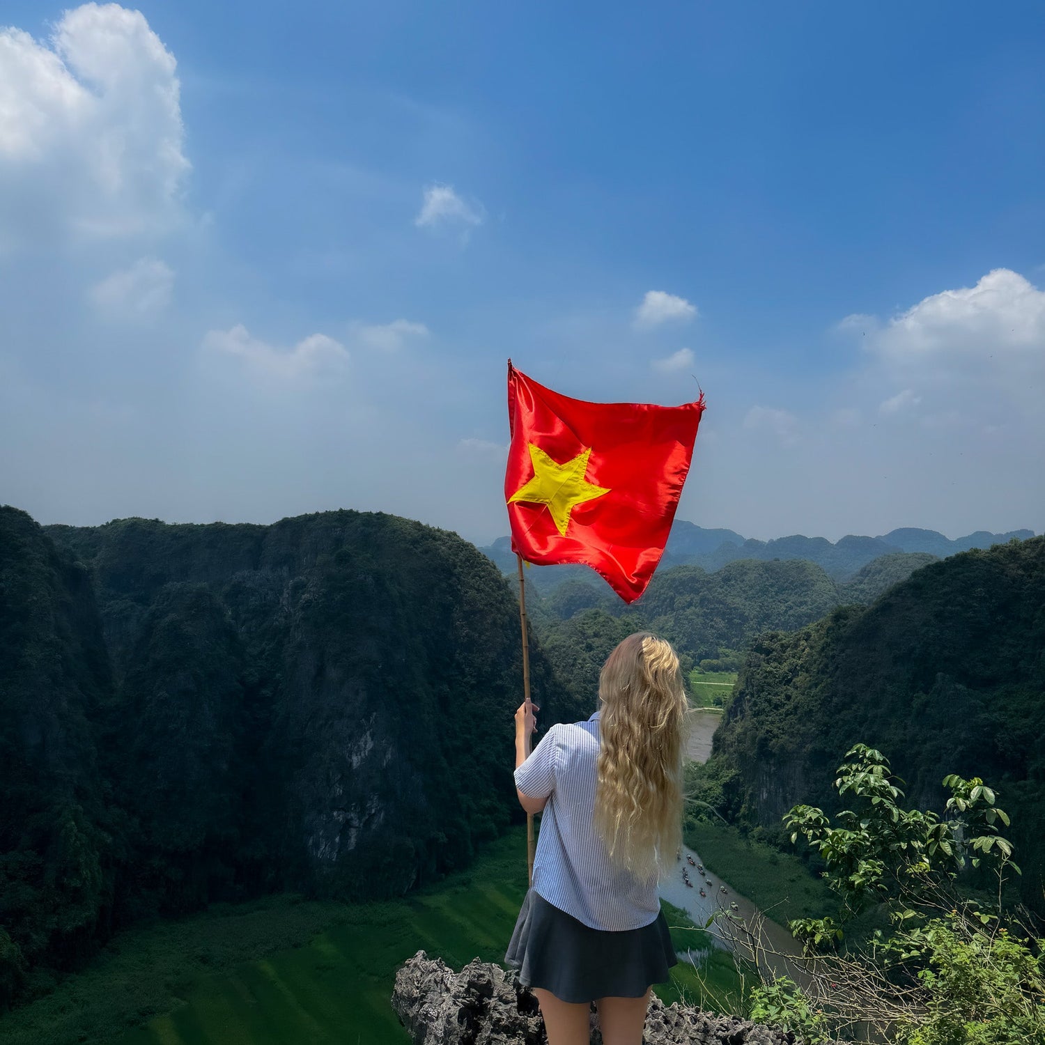 Woman standing on a mountain viewpoint holding the Vietnamese flag, overlooking green valleys under a bright blue sky – travel inspiration for elswre’s Vietnam jewelry collection.