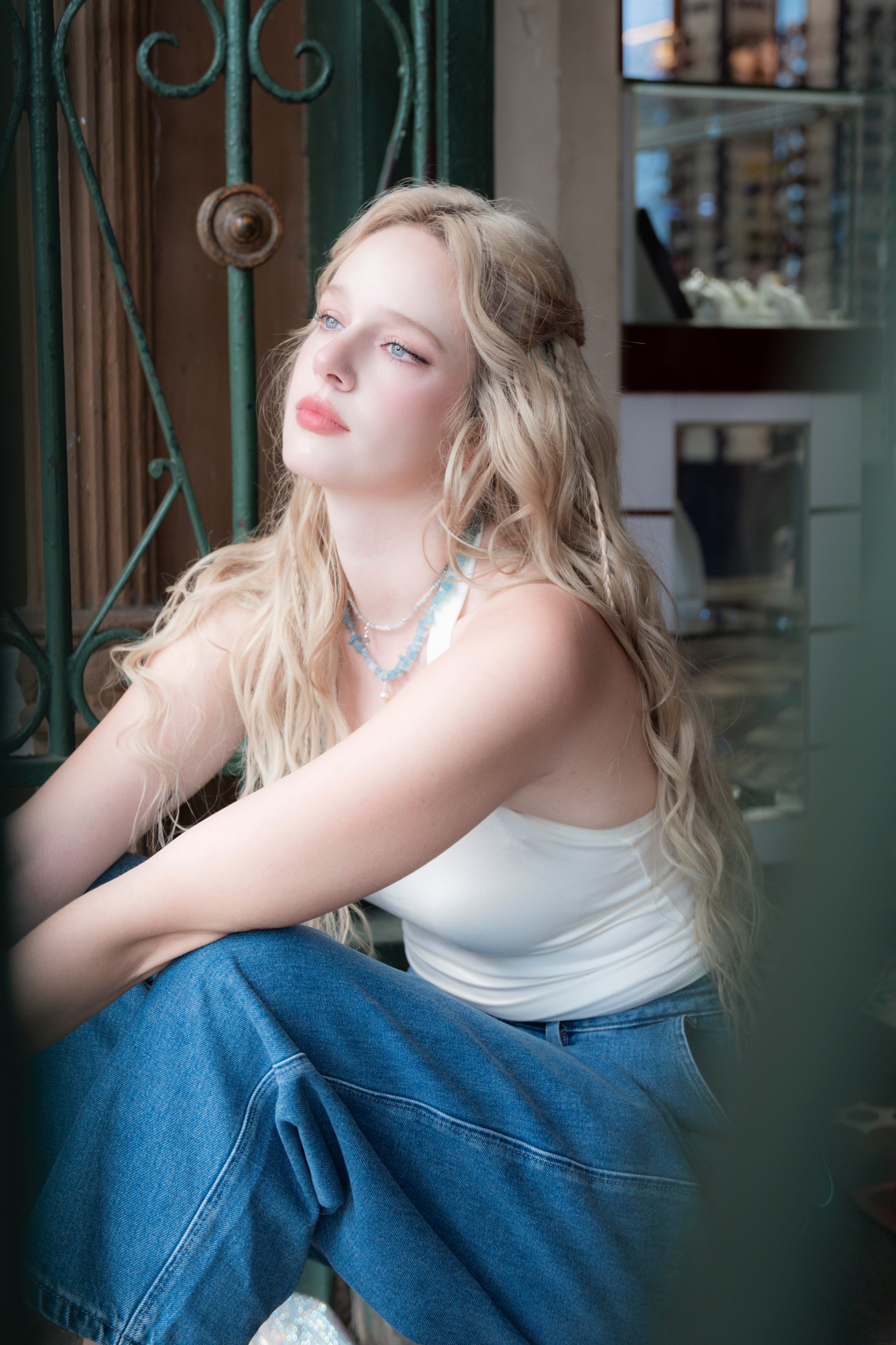 Woman wearing elswre’s aquamarine and pearl necklace, sitting near a green gate in soft natural light, wearing a white top and jeans.