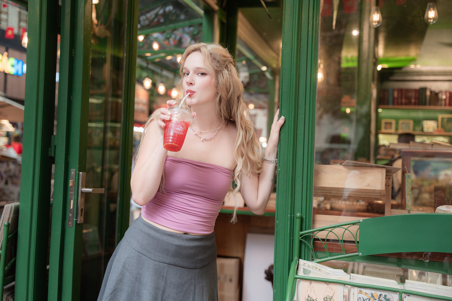 Woman wearing elswre jewelry, including a rose quartz and pearl necklace, enjoying a drink outside a café with green details in Saigon.