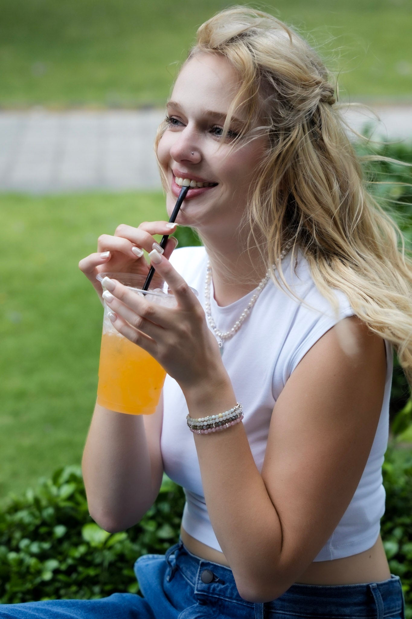 Woman wearing elswre pearl bracelet and necklace, smiling while drinking orange juice outdoors in a casual setting.