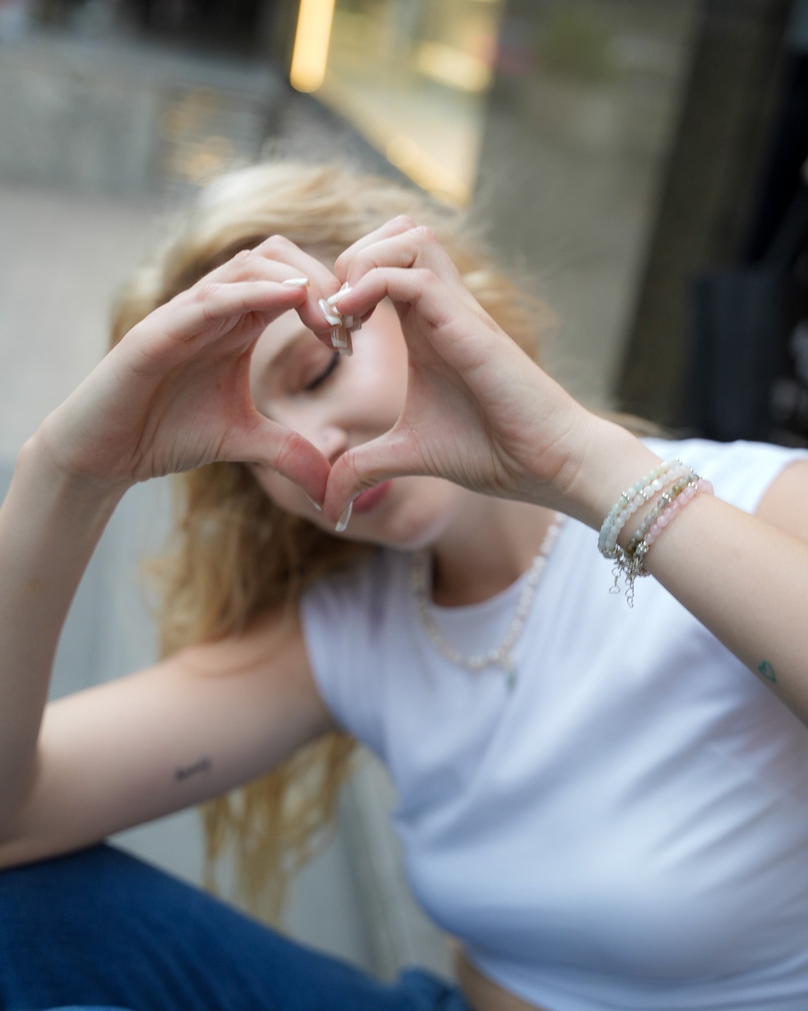Woman forming a heart with her hands while wearing layered gemstone and silver bracelets and a pearl necklace – casual, playful jewelry moment by elswre
