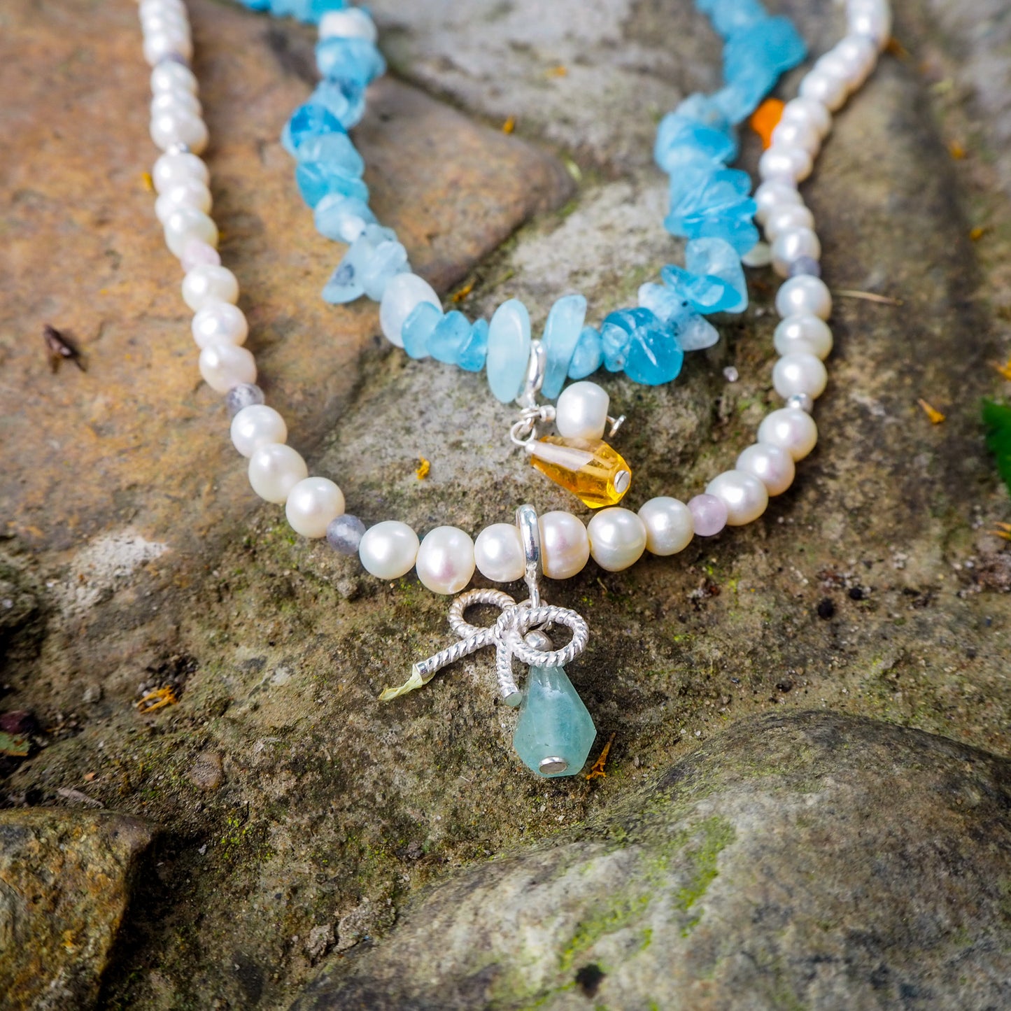 Pearl necklace with aquamarine and silver bow charms displayed on a natural moss-covered stone – handmade gemstone jewelry by elswre.