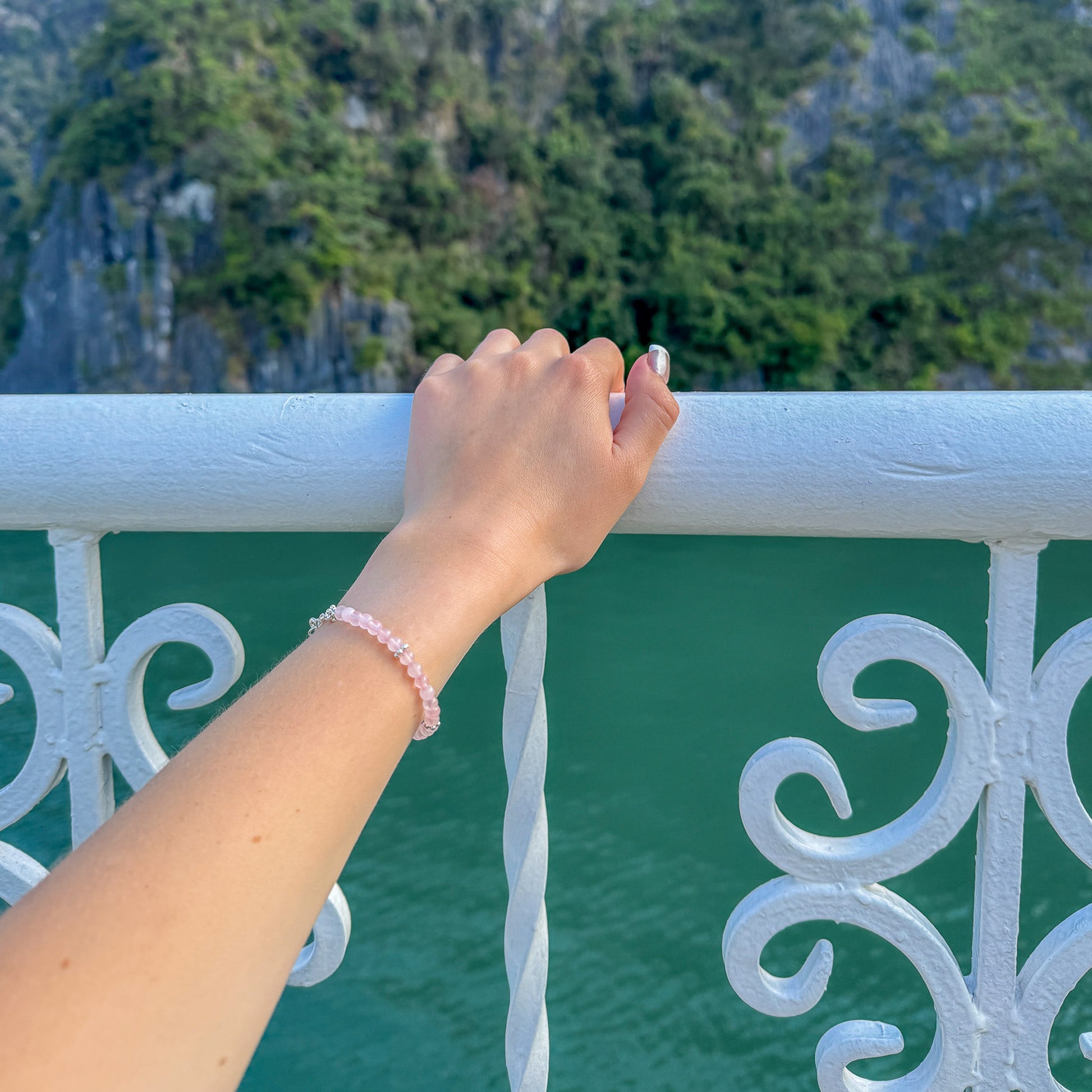 Hand wearing a rose quartz and silver bracelet resting on a white railing above turquoise water – travel-inspired handcrafted jewelry by elswre