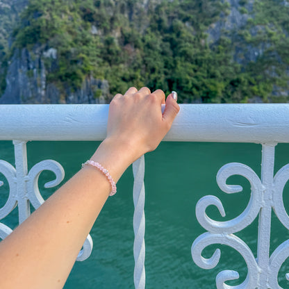 Hand wearing a rose quartz and silver bracelet resting on a white railing above turquoise water – travel-inspired handcrafted jewelry by elswre