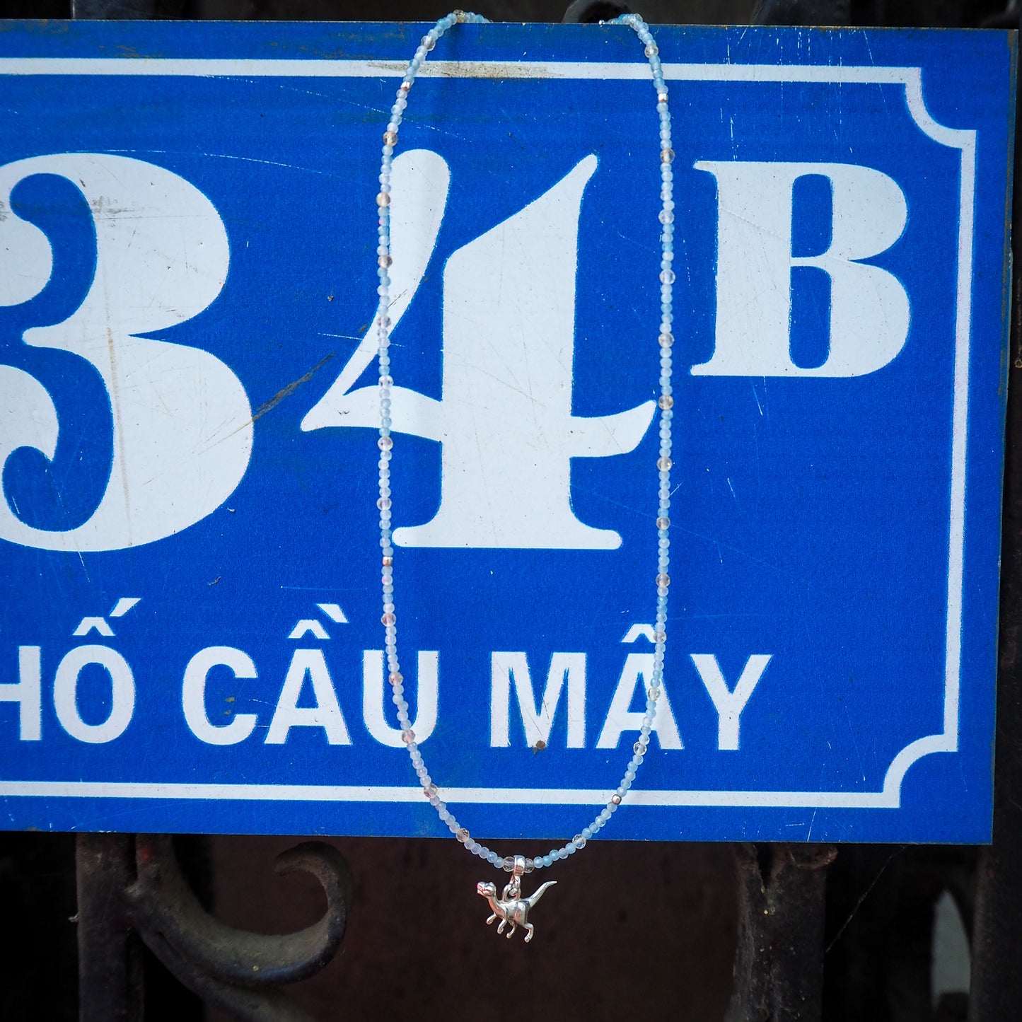 Aquamarine and light citrine beaded necklace with a small silver dinosaur charm hanging against a blue Vietnamese street sign – playful travel-inspired jewelry from elswre