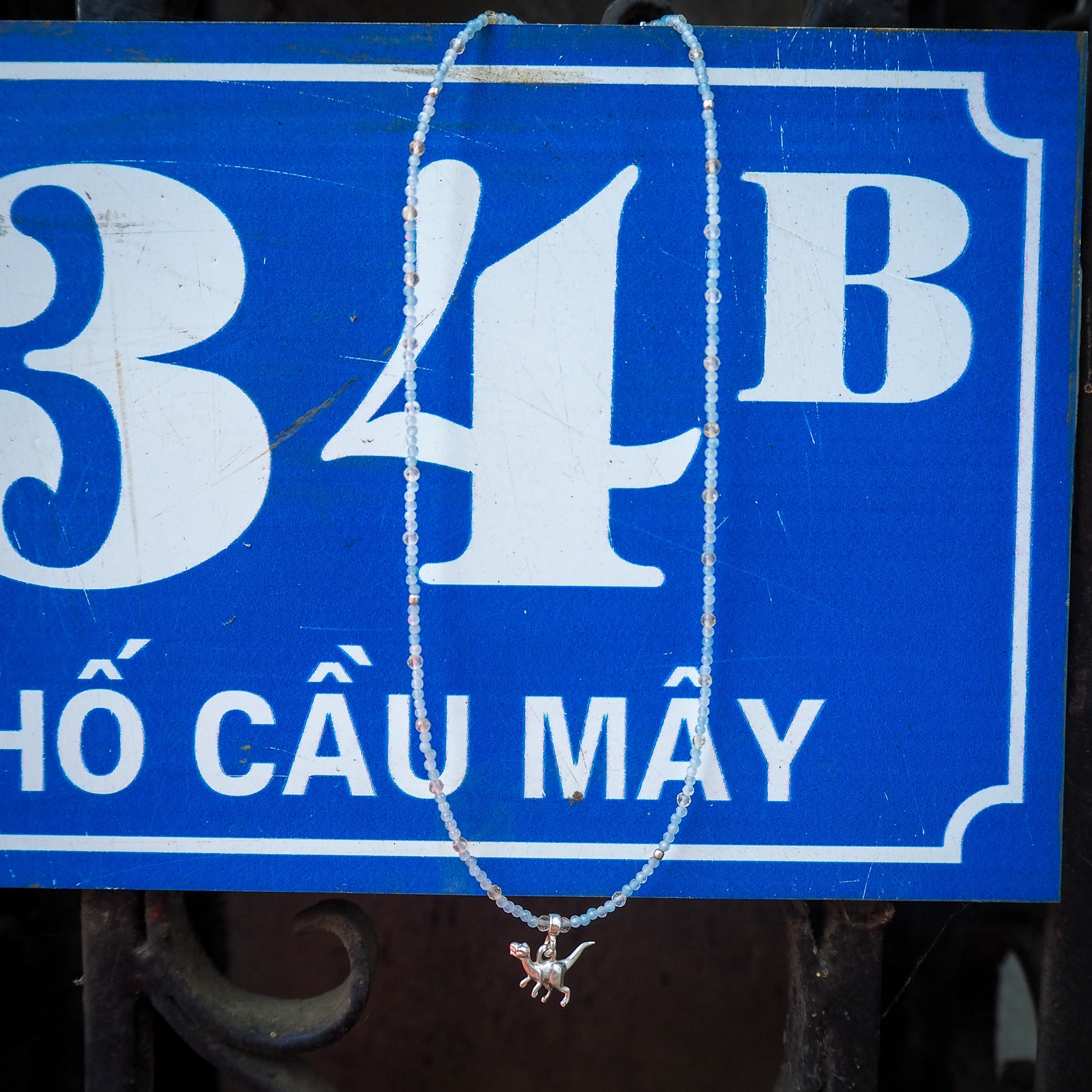 Aquamarine and light citrine beaded necklace with a small silver dinosaur charm hanging against a blue Vietnamese street sign – playful travel-inspired jewelry from elswre