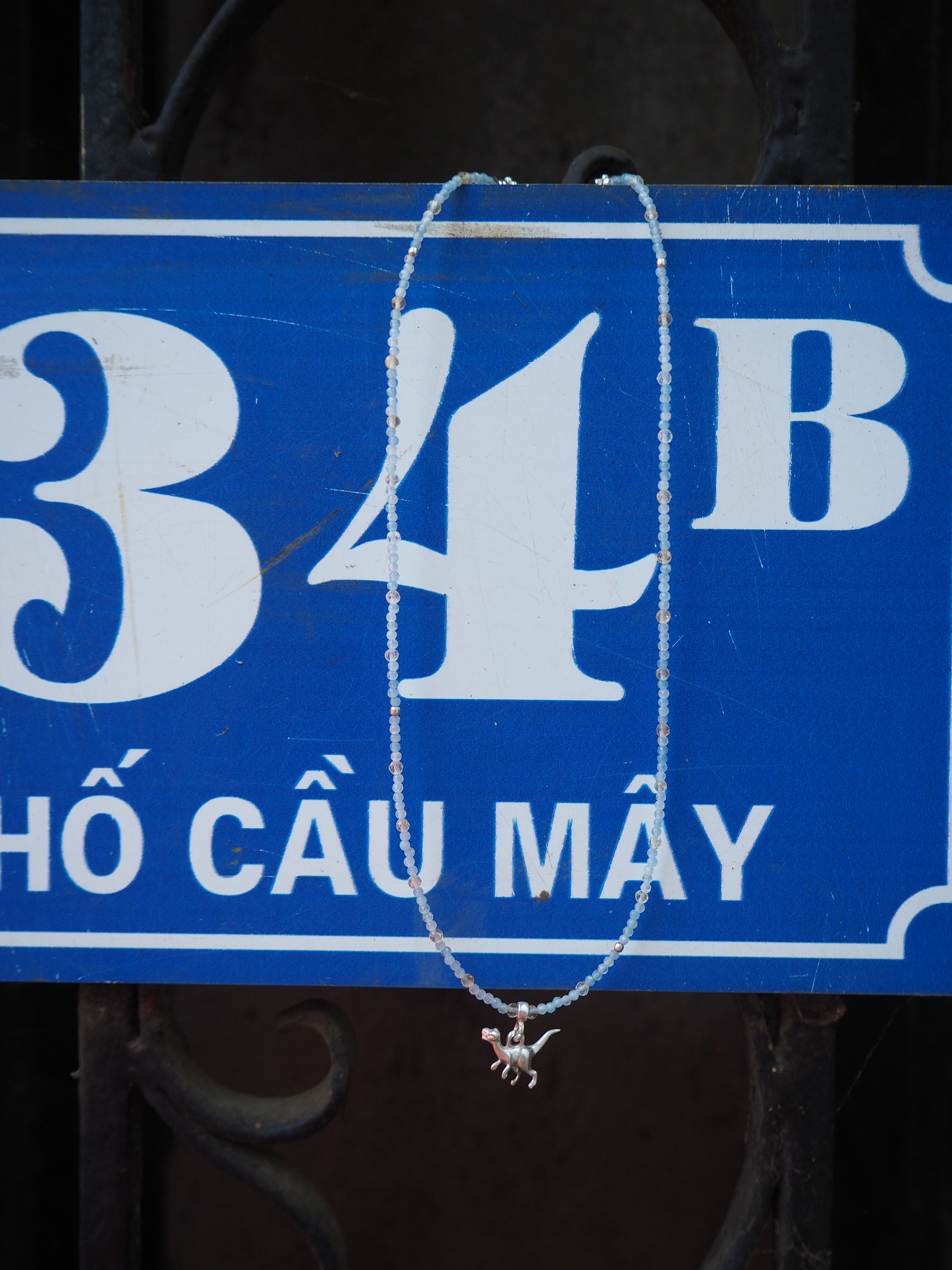 Silver beaded necklace with aquamarine and citrine charm displayed on a blue Vietnamese street sign, part of the elswre Da Nang jewelry collection.