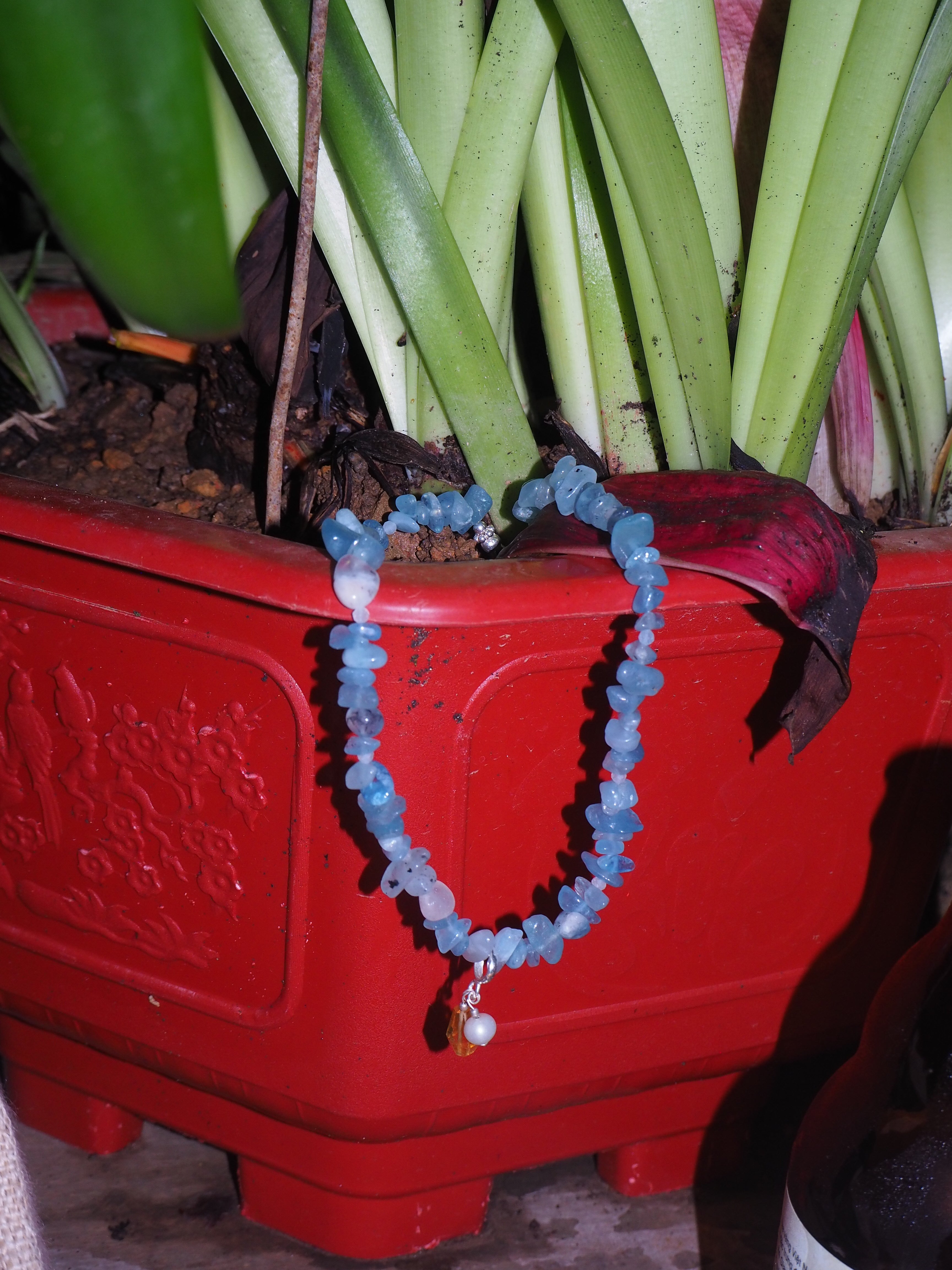 Aquamarine gemstone necklace with pearl and citrine charm by elswre jewelry, styled on a red flower pot with green tropical leaves in soft lighting.