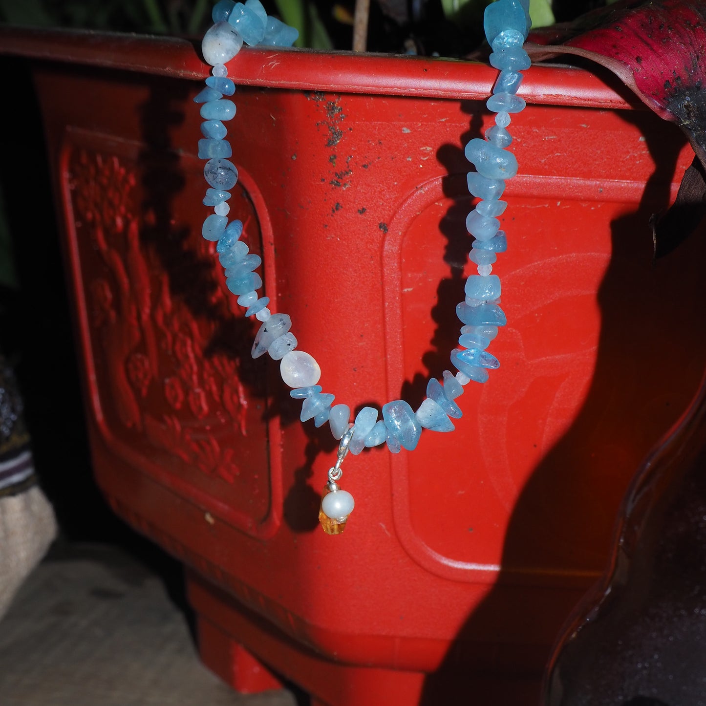 Aquamarine chip necklace with pearl and light citrine charm, displayed on a red planter – travel-inspired jewelry by elswre