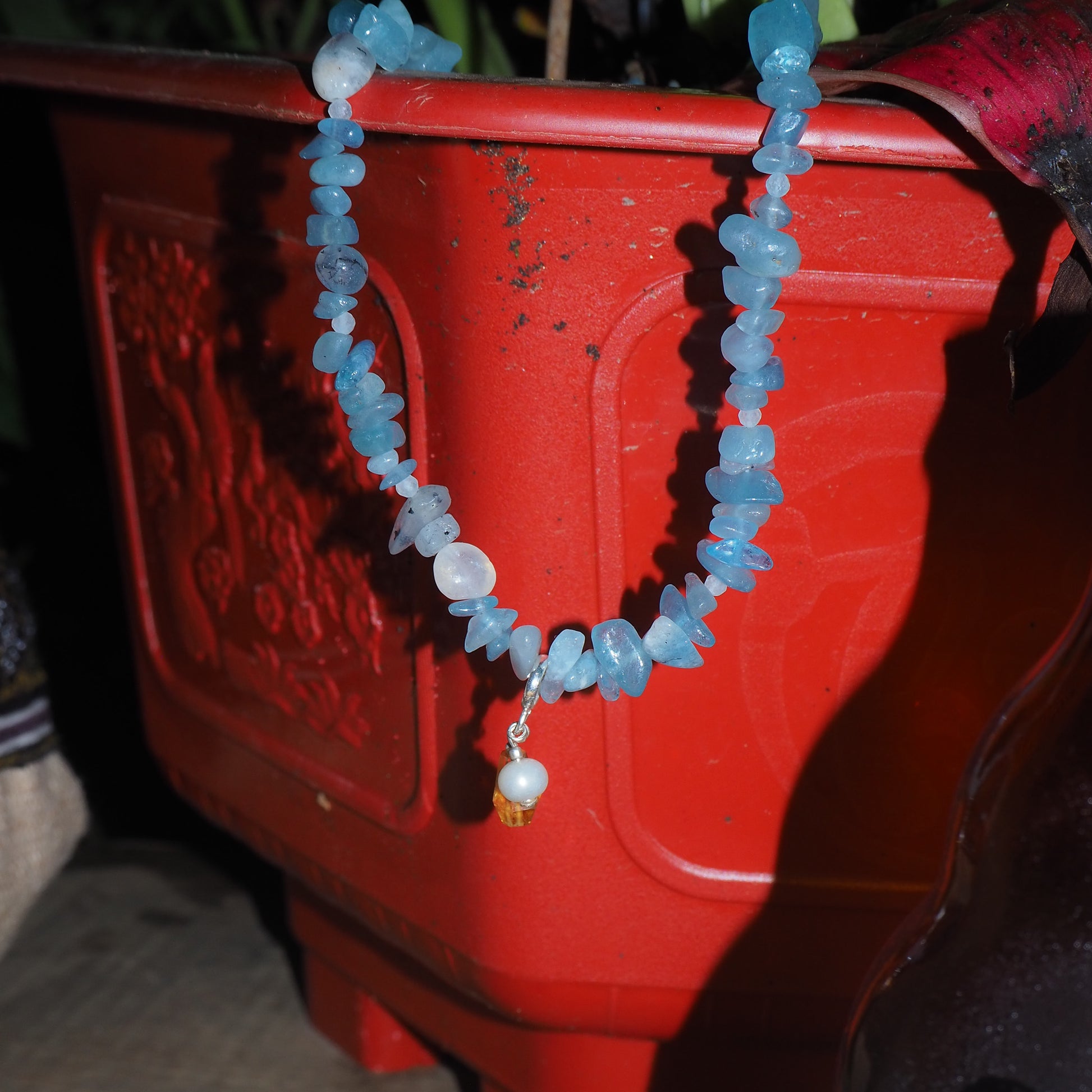 Aquamarine chip necklace with pearl and light citrine charm, displayed on a red planter – travel-inspired jewelry by elswre