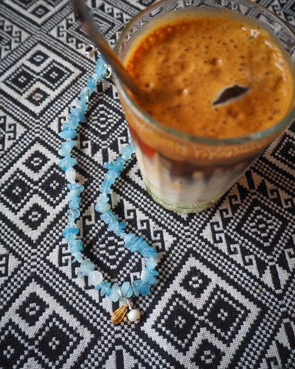 Aquamarine necklace with pearl and citrine charm placed beside an iced Vietnamese coffee on a patterned tablecloth – handmade gemstone jewelry by elswre